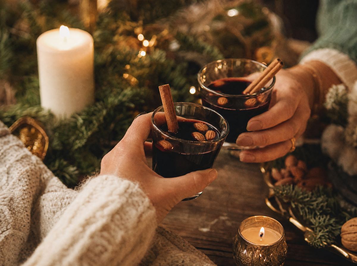Händer som håller glögg med kanelstänger intill levande ljus och granris